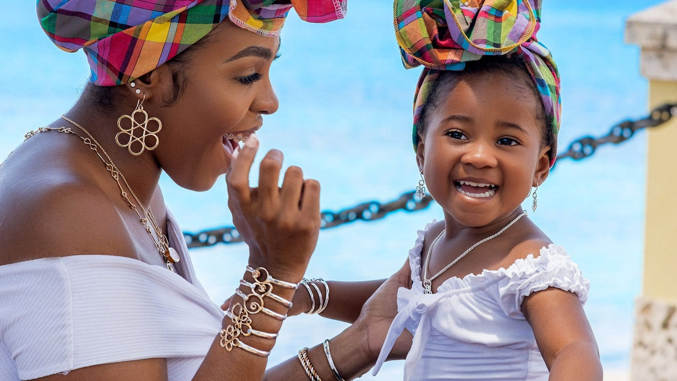 Woman and child wearing beautiful jewelry and colorful head wraps sitting on a bench with a caribbean sea