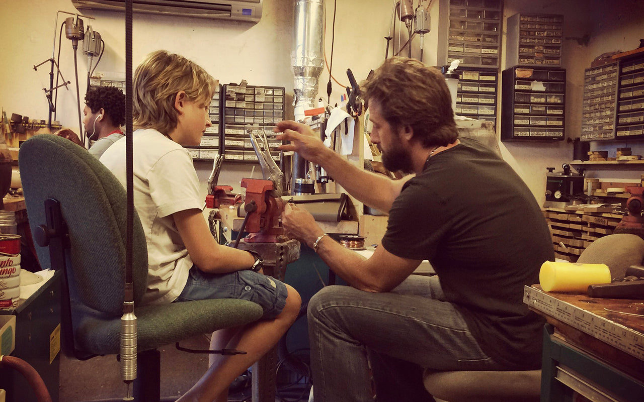 Two men in a workshop setting, one assisting the other with a task.